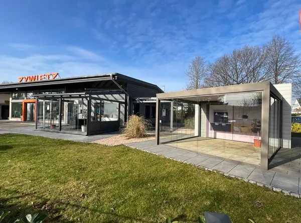 Moderner Wintergarten aus Glas und Aluminium vor einem Musterhaus bei sonnigem Wetter.