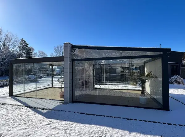Modernes Wintergarten-Glashaus mit klaren Glaswänden im verschneiten Garten bei Sonne.