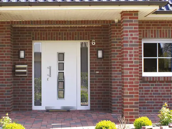 Moderne Haustür aus weißem Kunststoff in roter Klinkerfassade mit Seitenteil und Beleuchtung.